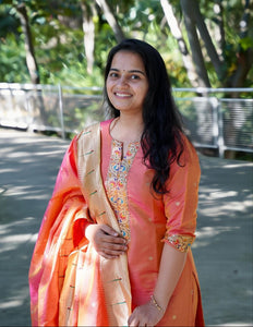 Woman in an orange traditional outfit standing outdoors with greenery in the background