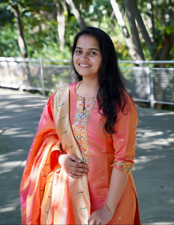 Woman in an orange traditional outfit standing outdoors with greenery in the background
